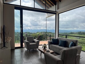 Una sala de estar moderna con un sofá, sillones y una mesa de centro. Amplias ventanas ofrecen vistas a un balcón y montañas.