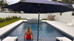 En swimmingpool med en person i vandet, en terrasse med stole og en parasol.
