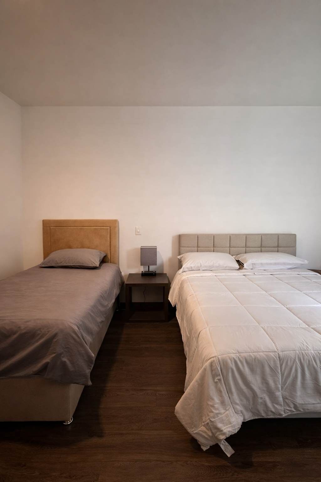 A bedroom with two beds, a nightstand with a lamp, and a wooden floor.