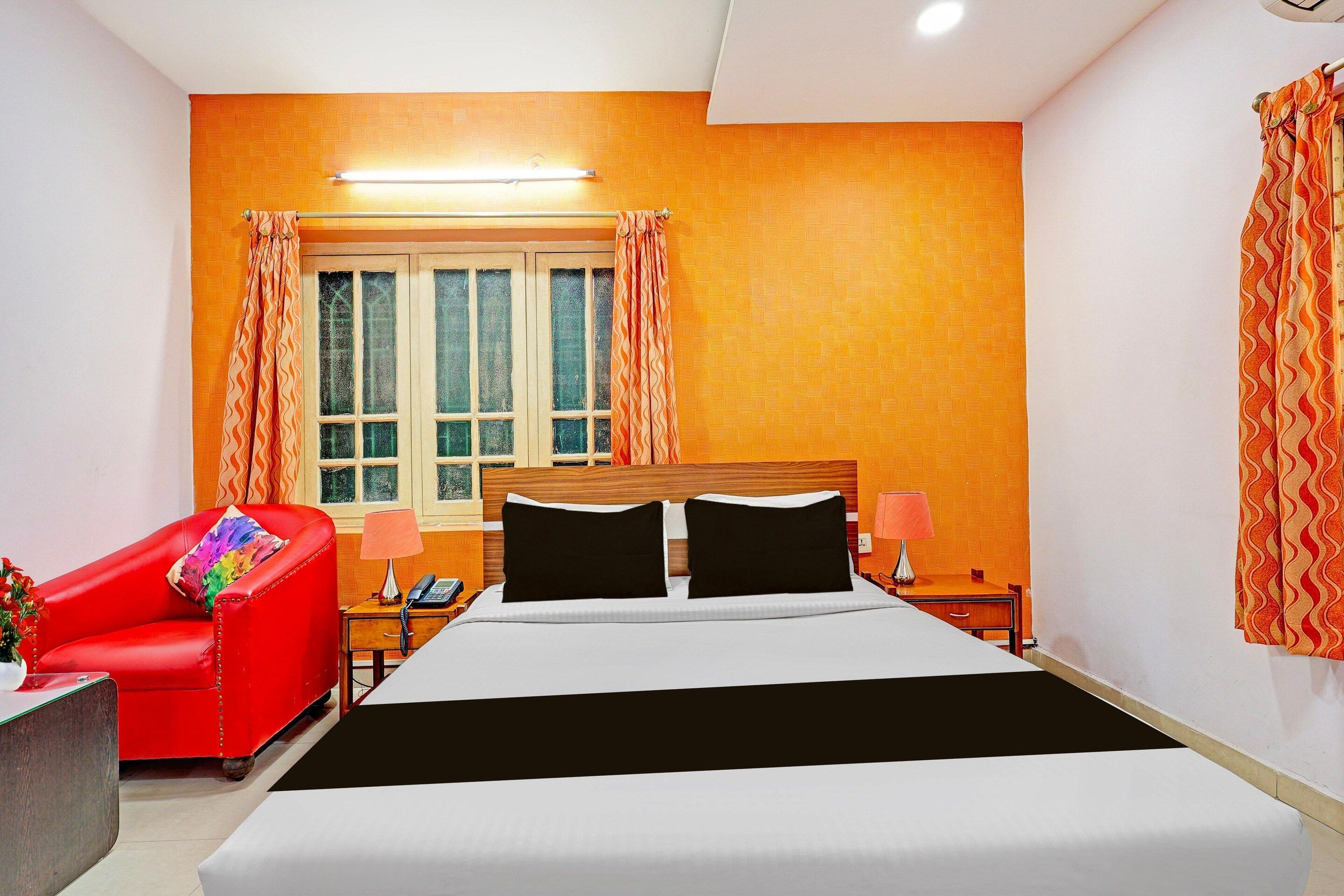 A hotel room with a bed, a red armchair, a nightstand, a lamp, and a window with curtains.