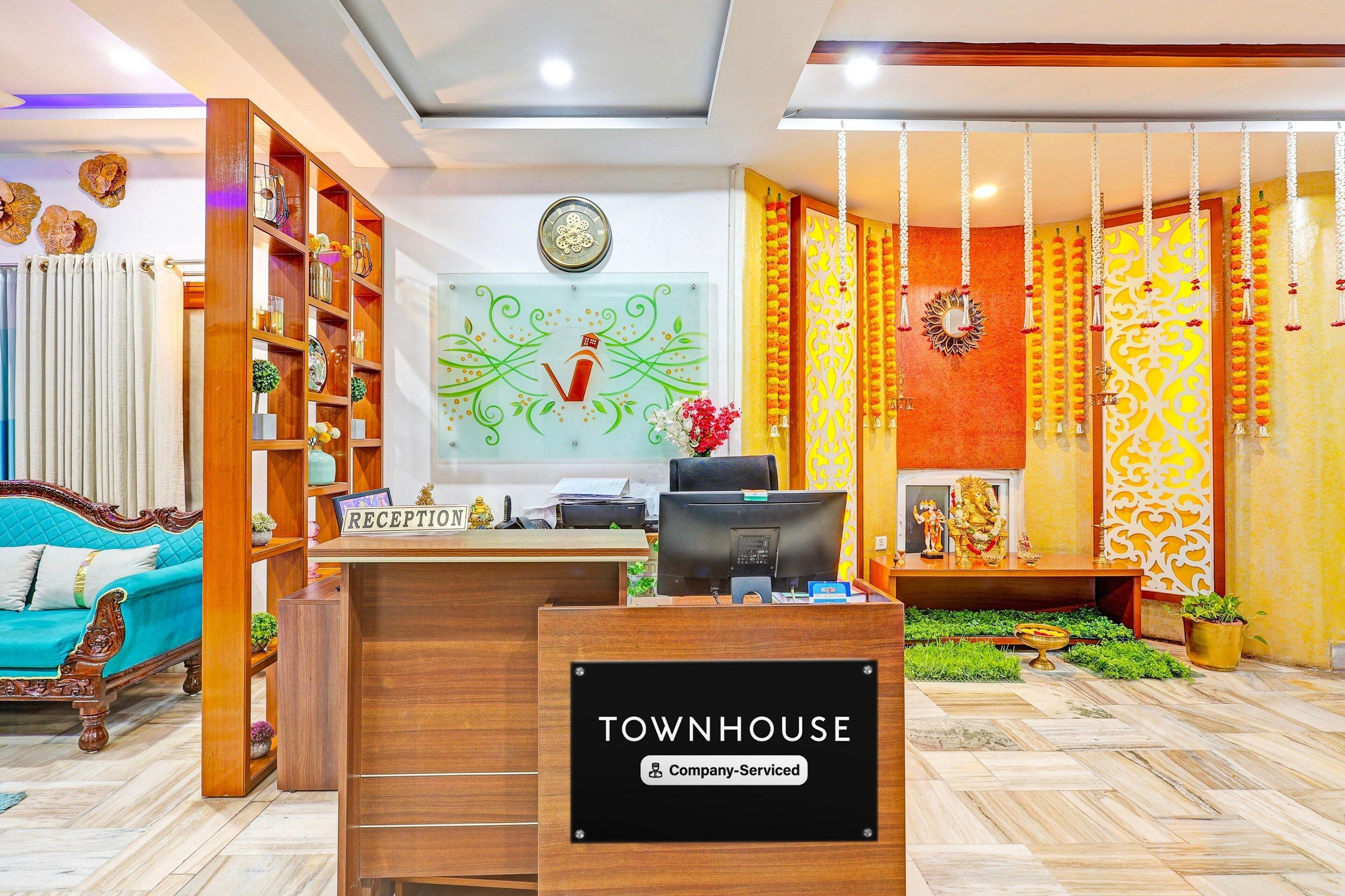A reception desk with a sign reading 'TOWNHOUSE Company Serviced,' a wooden bookshelf, a teal sofa, and a decorative wall with hanging ornaments.