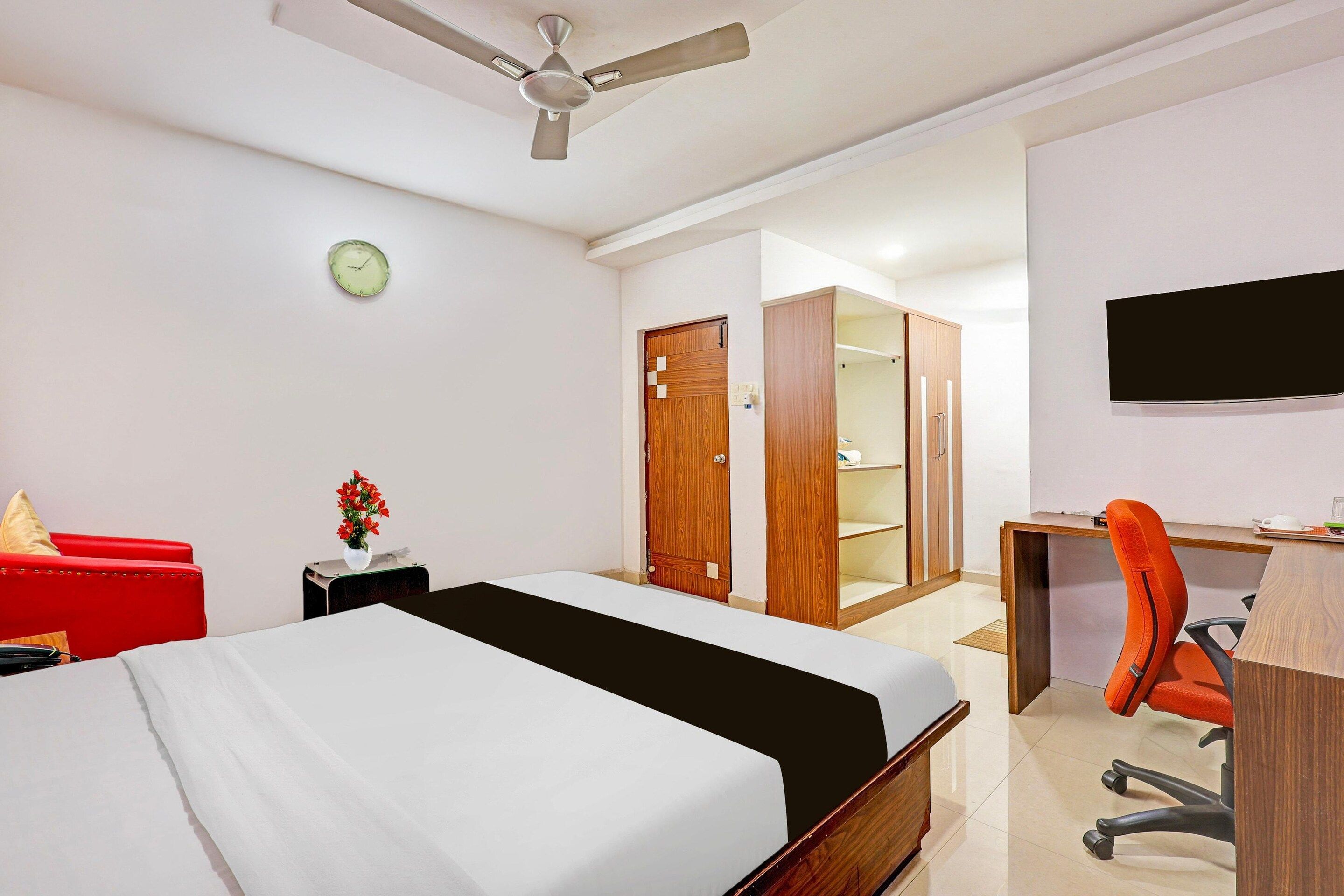 Ein Hotelzimmer mit einem Bett, einem Schreibtisch, einem orangefarbenen Bürostuhl, einem Deckenventilator, einer Uhr und einem an der Wand befestigten Fernseher.