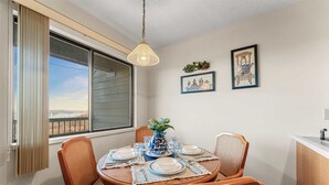 Un coin repas avec une table ronde, quatre chaises, un vase de fleurs et une vue sur une étendue d’eau, visible à travers une grande fenêtre.