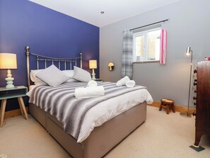 A neatly made bed with pillows and a rolled towel, a bedside table with a lamp, a window with curtains, and a small stool. - The Green (Ripon)