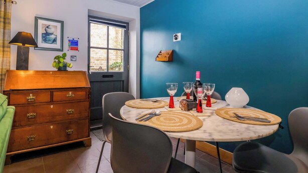A dining area with a round table, chairs, a wooden chest of drawers, a window, and a blue wall. - The Green (Ripon)