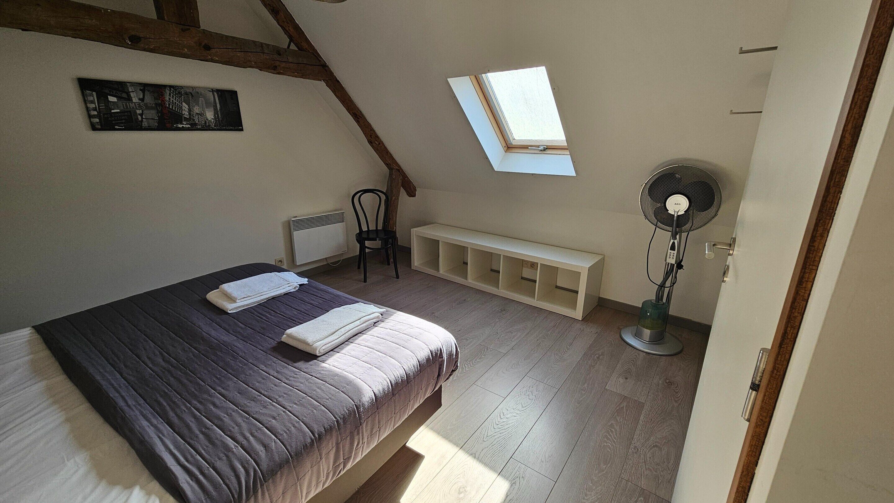 A bedroom with a bed, a bench, a radiator, a fan, and a skylight.