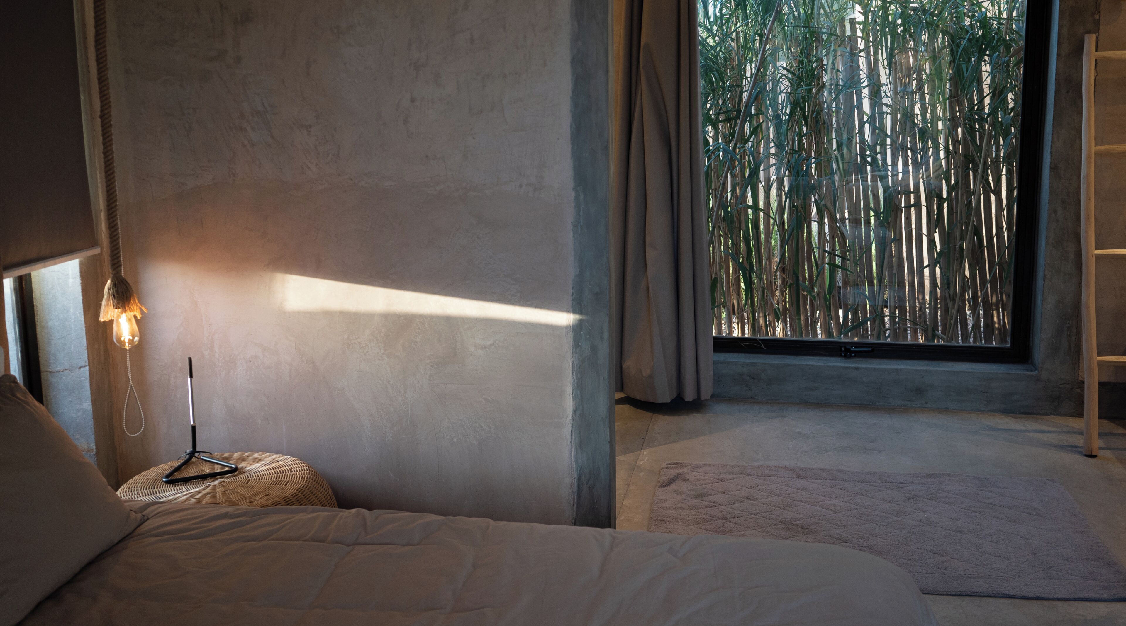 A bedroom with a bed, a bedside lamp, a window with curtains, and a view of bamboo outside.