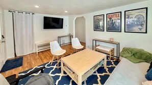 A modern living room with a TV, two white chairs, a wooden table, and framed cityscape pictures on the wall. - NEW! Peaceful Bungalow • Close to Lightrail • Yard (Seattle)