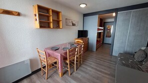 Ein Essbereich mit einem Tisch für vier Personen, Holzsesseln, einem Holzschrank und einem Fernseher auf einem Standfuß.