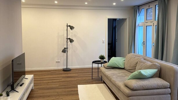 Una sala de estar moderna con un sofá, un televisor, una lámpara de pie y un ventanal con cortinas.