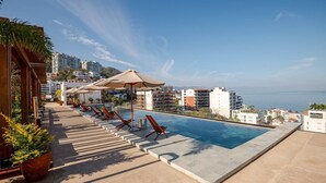 Ein Poolbereich mit Liegestühlen, Sonnenschirmen und Blick auf das Meer und Gebäude.