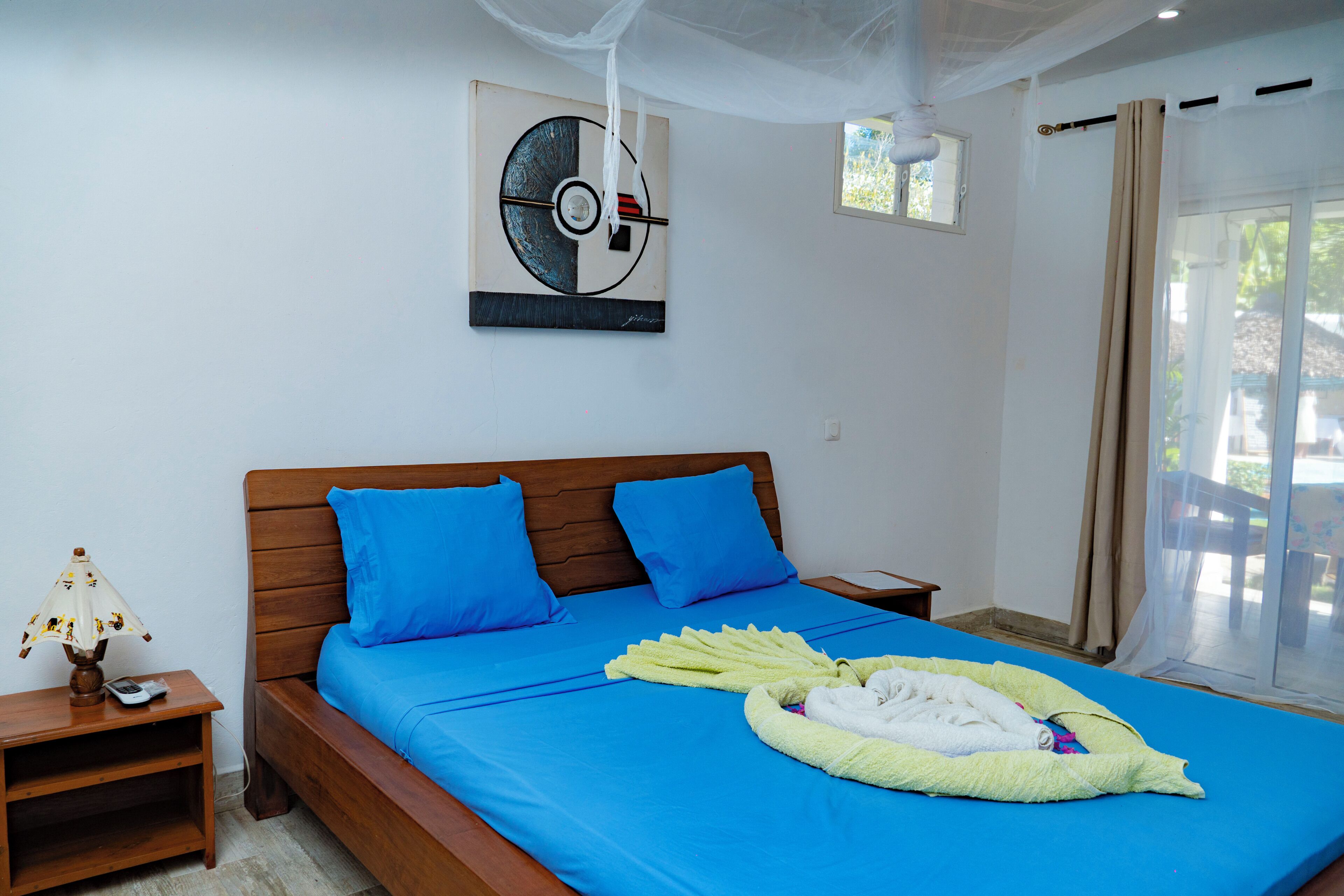 A bedroom with a bed, blue pillows, a bedside table with a lamp, a wall clock, and a window with curtains.