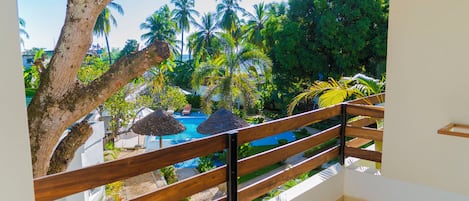 A balcony with a wooden railing overlooking a swimming pool, surrounded by lush greenery and palm trees.