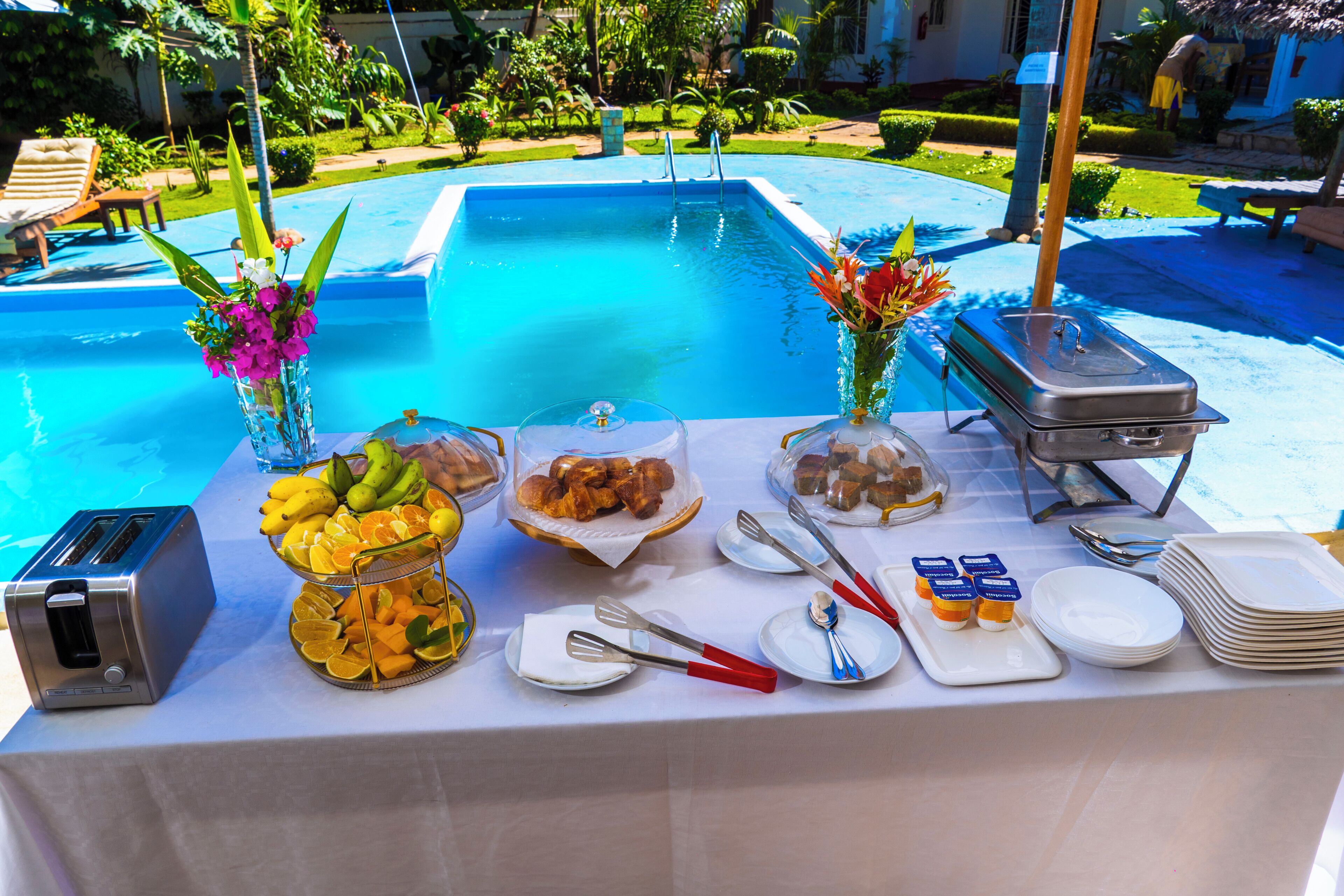 A buffet table with food, utensils, and a toaster by a poolside.