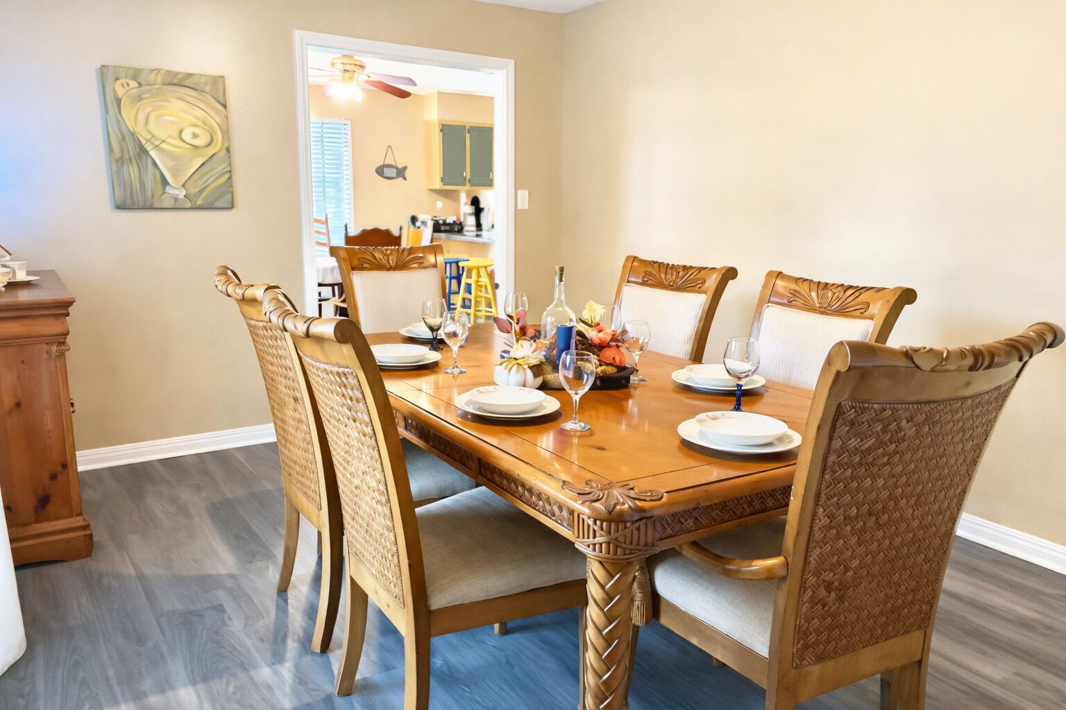 Une salle à manger avec une table en bois et des chaises en osier, un tableau accroché au mur et une cuisine visible à travers une porte ouverte.