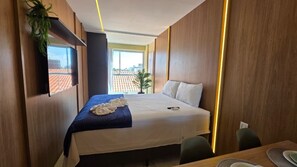 Ein Hotelzimmer mit Bett, Wandfernseher, Schreibtisch mit Stuhl und Blick auf die Stadtlandschaft durch das Fenster.