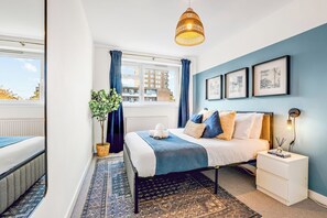 A bedroom with a bed, bedside table, lamp, and a view of buildings through a window. - Spacious 2-Bed Apartment In Hackney (Greater London)