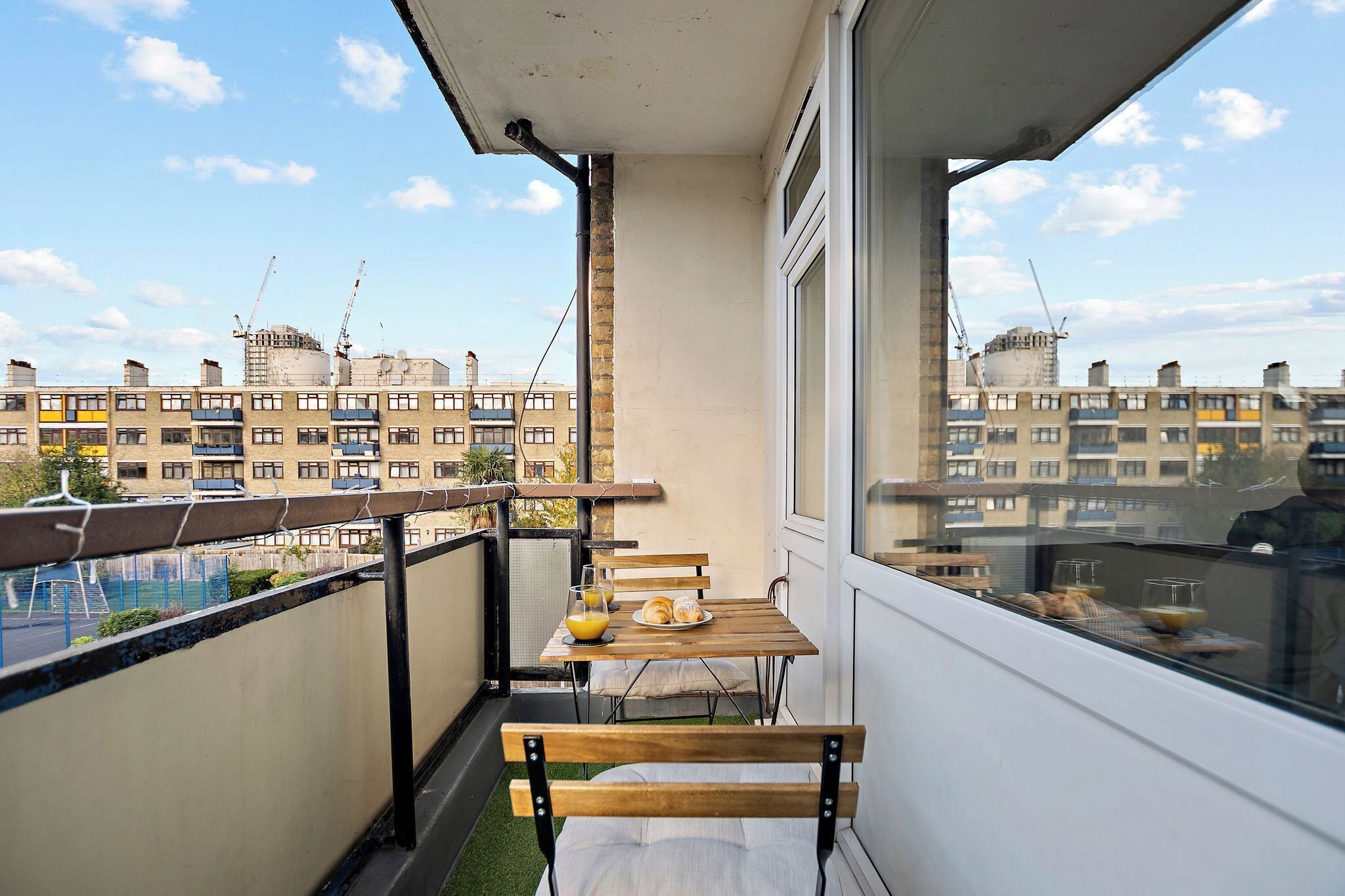 A balcony with a wooden table and chairs, a bench, and a view of a cityscape with buildings and construction cranes.