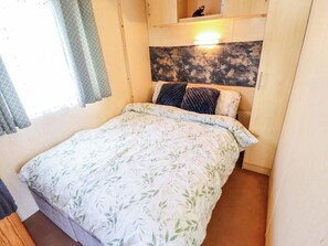 A bedroom with a bed, a headboard with a light fixture, a window with curtains, and a small shelf above the bed. - 517 - Promenade Caravan Site (Skegness)