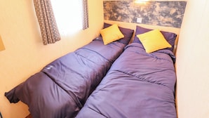 A small, single-bed room with a headboard, a window with curtains, and a wall-mounted light. - 517 - Promenade Caravan Site (Skegness)