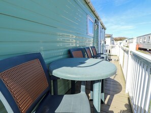 A covered outdoor seating area with a table and chairs, a white railing, and a view of a row of mobile homes in the background. - 517 - Promenade Caravan Site (Skegness)