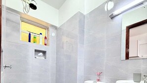 Et badeværelse med toilet, håndvask, bruser og en lille hylde med toiletartikler.