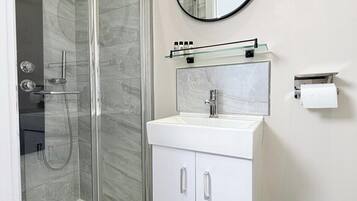 Un baño moderno con cabina de ducha de vidrio, un lavabo blanco con espejo superior y un inodoro.