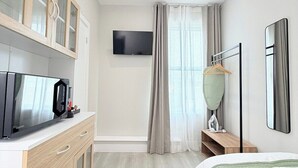 Habitación de hotel moderna con televisión de pantalla plana, cama con ropa de cama verde, aparador de madera con puertas de cristal, pequeño escritorio con lámpara y ventana grande con cortinas.