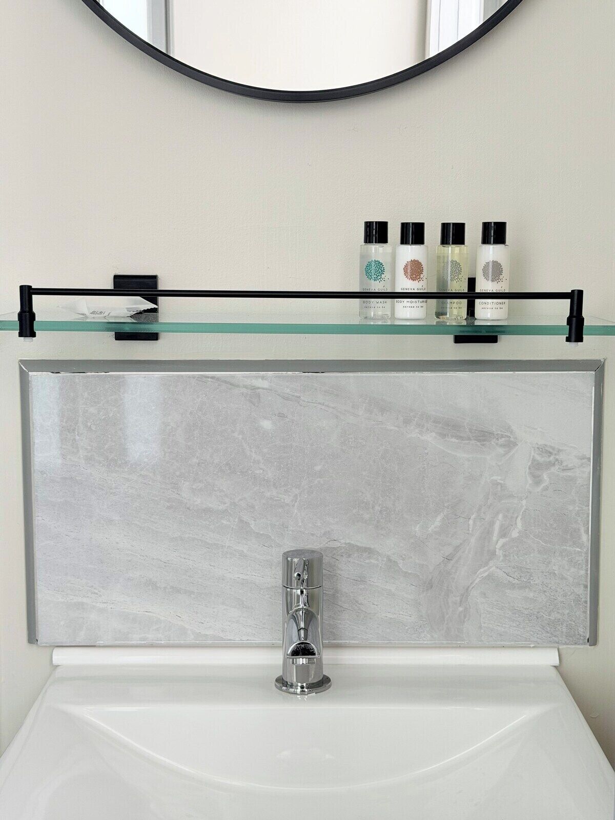 Un bagno con lavandino in marmo, un rubinetto cromato e uno scaffale in vetro sopra lo specchio, con prodotti per l'igiene personale.