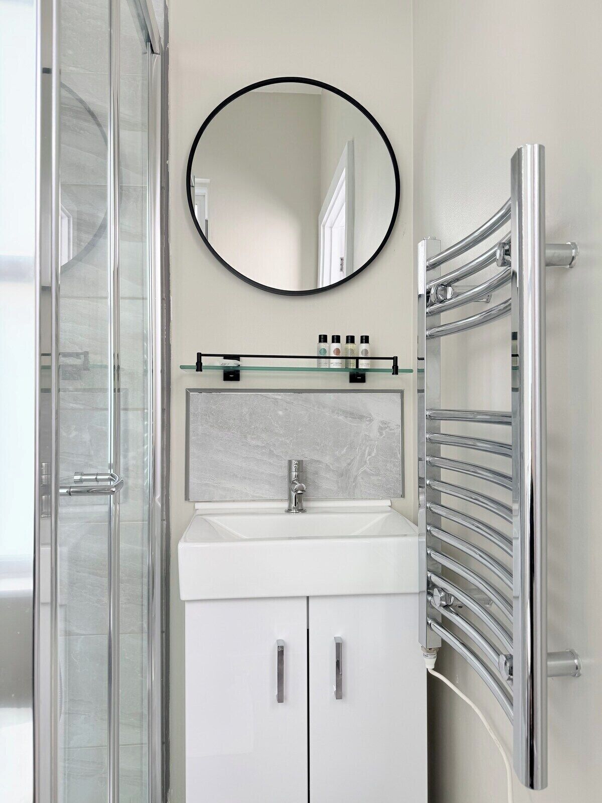 A bathroom with a glass shower enclosure, a white sink with a marble backsplash, a round mirror, and a chrome towel rack.