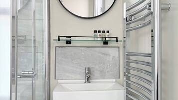 A bathroom with a glass shower enclosure, a white sink with a marble backsplash, a round mirror, and a chrome towel rack.