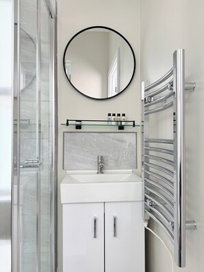 Een badkamer met een glazen douchecabine, een wit wastafelmeubel met een marmeren achterwand, een ronde spiegel en een chroomhanddoekhouder.