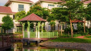 Um gazebo com telhado vermelho e grades verdes, um lago com ponte de madeira, e uma fileira de casas com telhados vermelhos e molduras de janelas verdes.