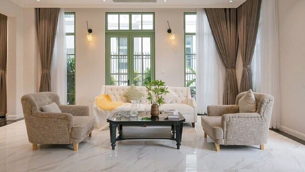 Uma sala de estar ampla com um sofá branco, duas poltronas, piso de mármore e grandes janelas com molduras verdes.