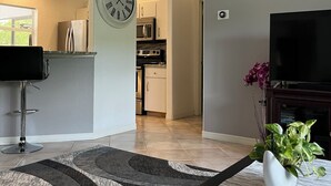 Ett modernt vardagsrum med en platt-TV, en klocka, en matplats och ett kök i bakgrunden.