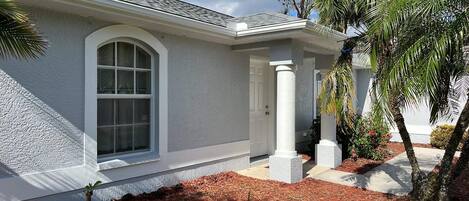 Una casa con un ventanal arqueado grande, columnas blancas y un jardín ajardinado con mantillo y palmeras.