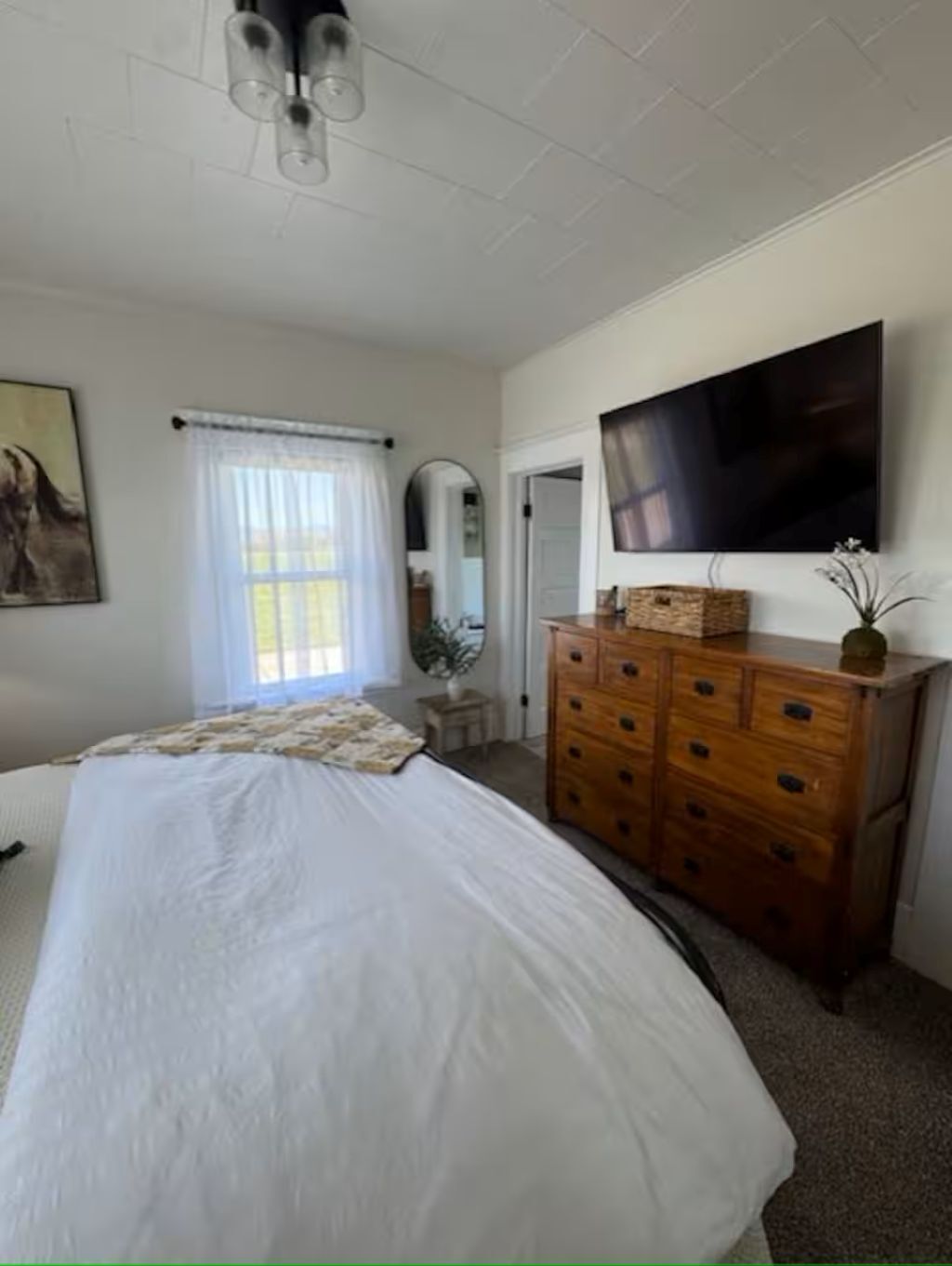 Ein Schlafzimmer mit einem Bett, einer Holzkommode, einem Flachbildfernseher und einem Fenster mit Vorhängen.