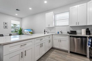 A modern kitchen with white cabinetry, a stainless steel dishwasher, and a countertop with a wine glass and a picture frame. - Stylish Newly Built Retreat (West Palm Beach)