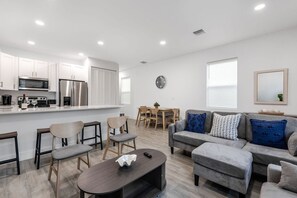 A modern kitchen with white cabinets, a dining area with a wooden table and chairs, and a living room with a grey sofa and ottoman. - Stylish Newly Built Retreat (West Palm Beach)