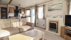 A caravan interior with a sofa, table, kitchen area, and a fireplace. - 23 Gull Court (Porthmadog)