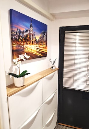 Una habitación con una pintura de paisaje urbano, una orquídea en maceta, un estante de madera, un ventanal con marco negro y un armario blanco.