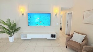 Sala de estar con un televisor de pantalla plana montado en la pared, un mueble blanco debajo, dos lámparas de pared, una planta en maceta y un sofá marrón con un cojín blanco.