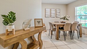Comedor con mesa de madera, sillas, planta en maceta, cuadros enmarcados y una ventana con persianas.