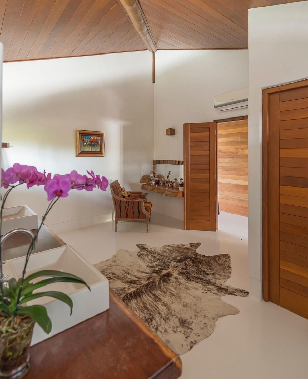 A modern interior with a wooden ceiling, a dining area with a chair, a small table, a mirror, a shelf with toiletries, and a large rug.