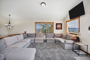 A living room with a sectional sofa, a flat-screen TV, a chandelier, and a window with blinds.