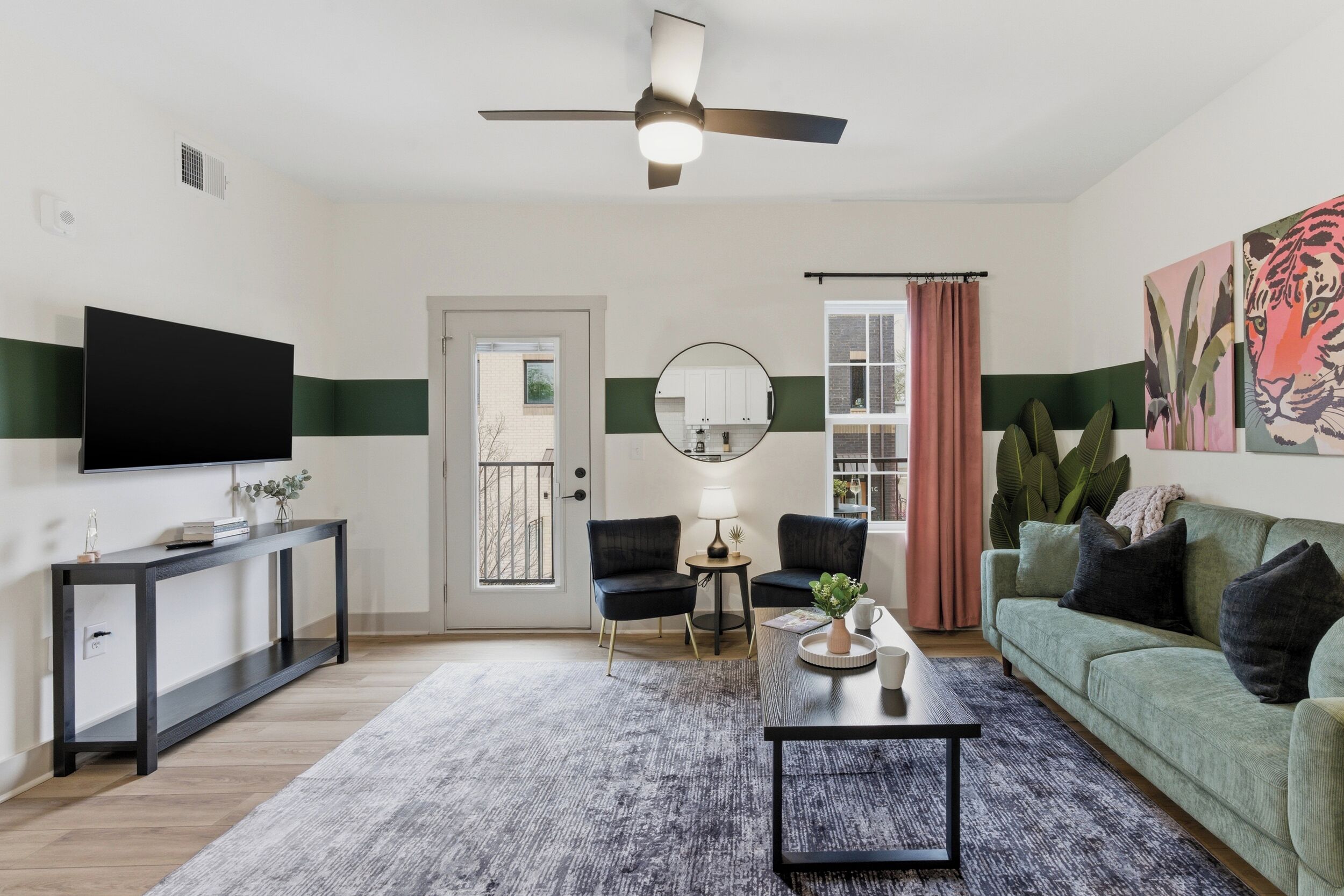 Un salon équipé d’un téléviseur à écran plat, d’un ventilateur de plafond, d’un miroir rond, d’un canapé vert et d’une table basse.