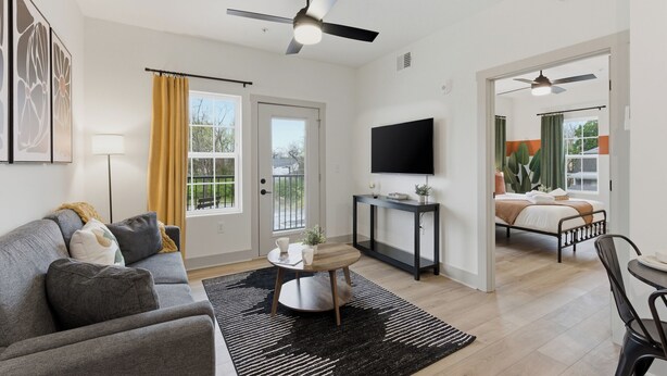 A modern living room with a sofa, a coffee table, a TV, and a ceiling fan. - The Lofts On Maney (Murfreesboro)