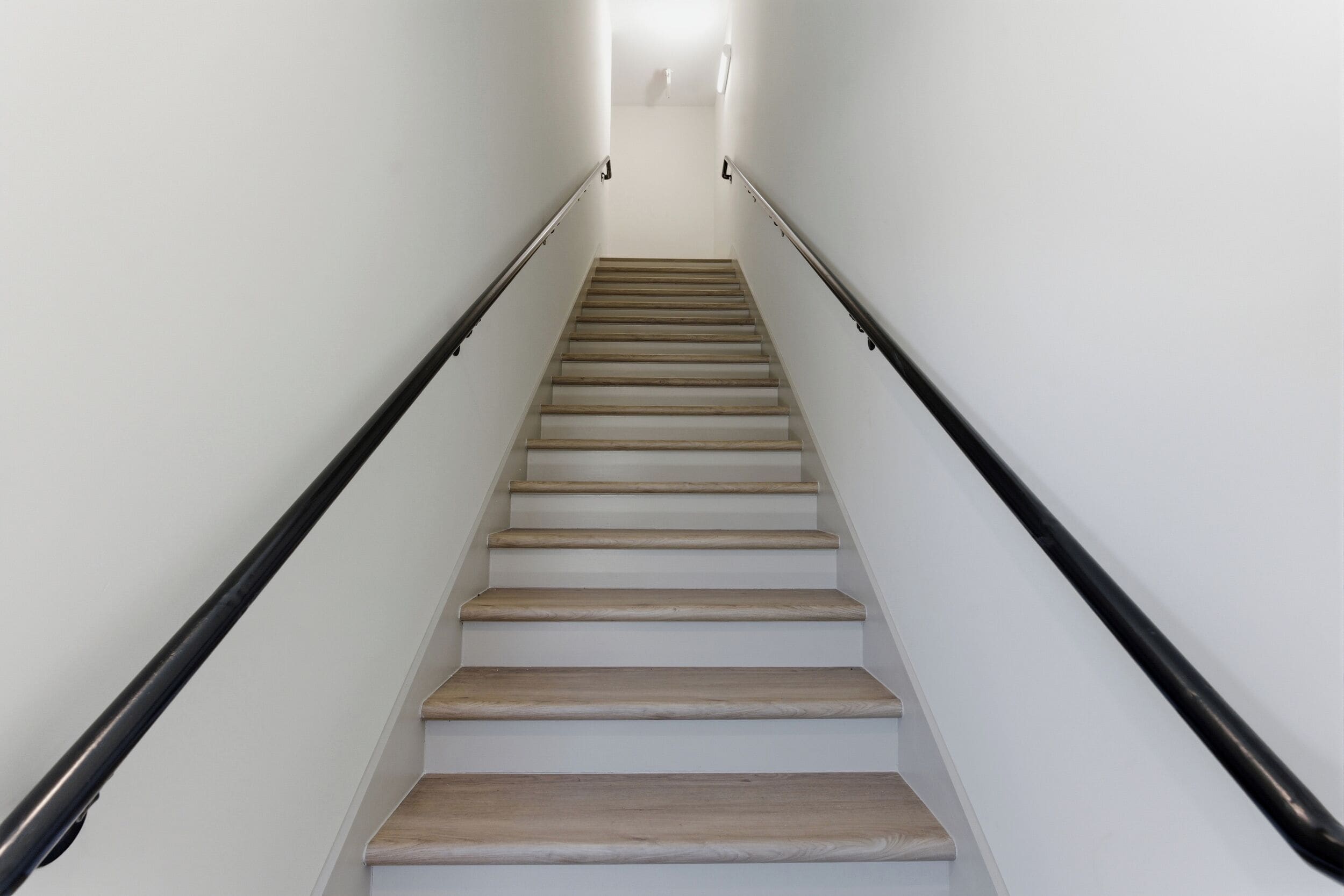 Una scala con gradini in legno e corrimano neri, che porta a un pianerottolo illuminato da un lampadario.