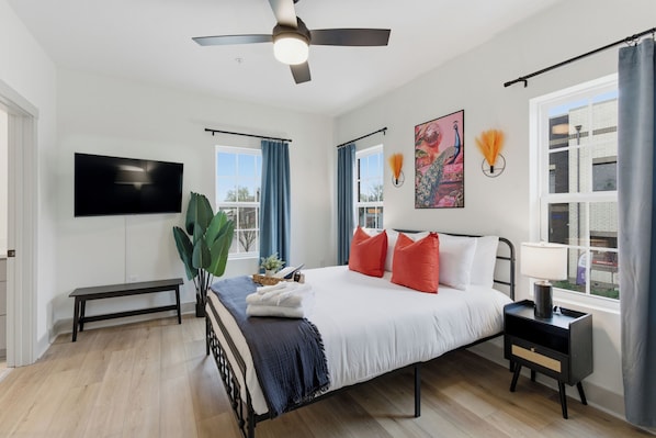 A bedroom with a bed, a TV, a ceiling fan, a window with curtains, and a plant.