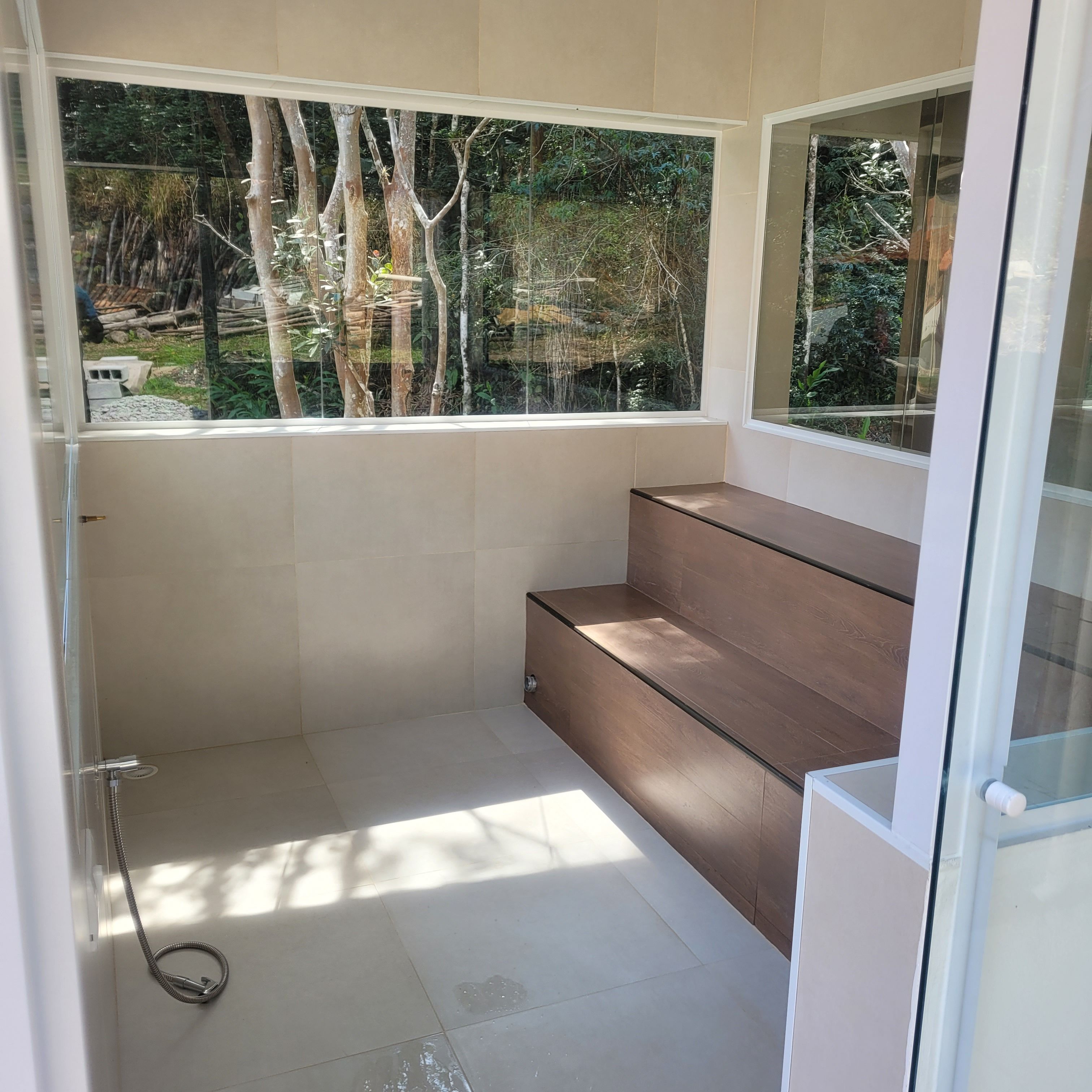 A modern bathroom with a glass shower enclosure, a wooden bench, and a view of a forest through a large window.